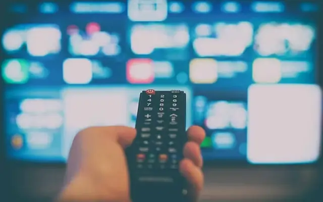 Mão segurando o controle remoto diante da TV